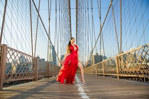 Woman in flowing red dress posing on Brooklyn Bridge walkway with NYC skyline in background.