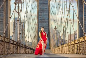 Woman in red dress posing on Brooklyn Bridge with city skyline in background.