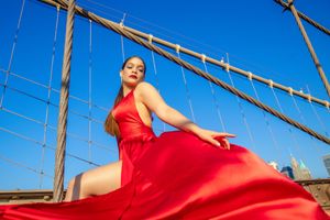 Woman in flowing red dress posing on Brooklyn Bridge with blue sky backdrop.