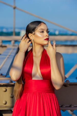 Woman in red satin deep-V halter dress poses on bridge, hands near face, red lips, sleek hair, blue sky background.