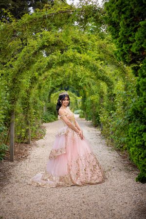 Young woman in pink gold quinceañera gown poses under lush green garden archway on gravel path.