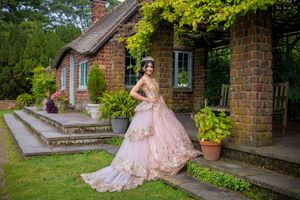 Young woman in elegant pink gold ball gown poses in lush garden beside charming brick cottage.