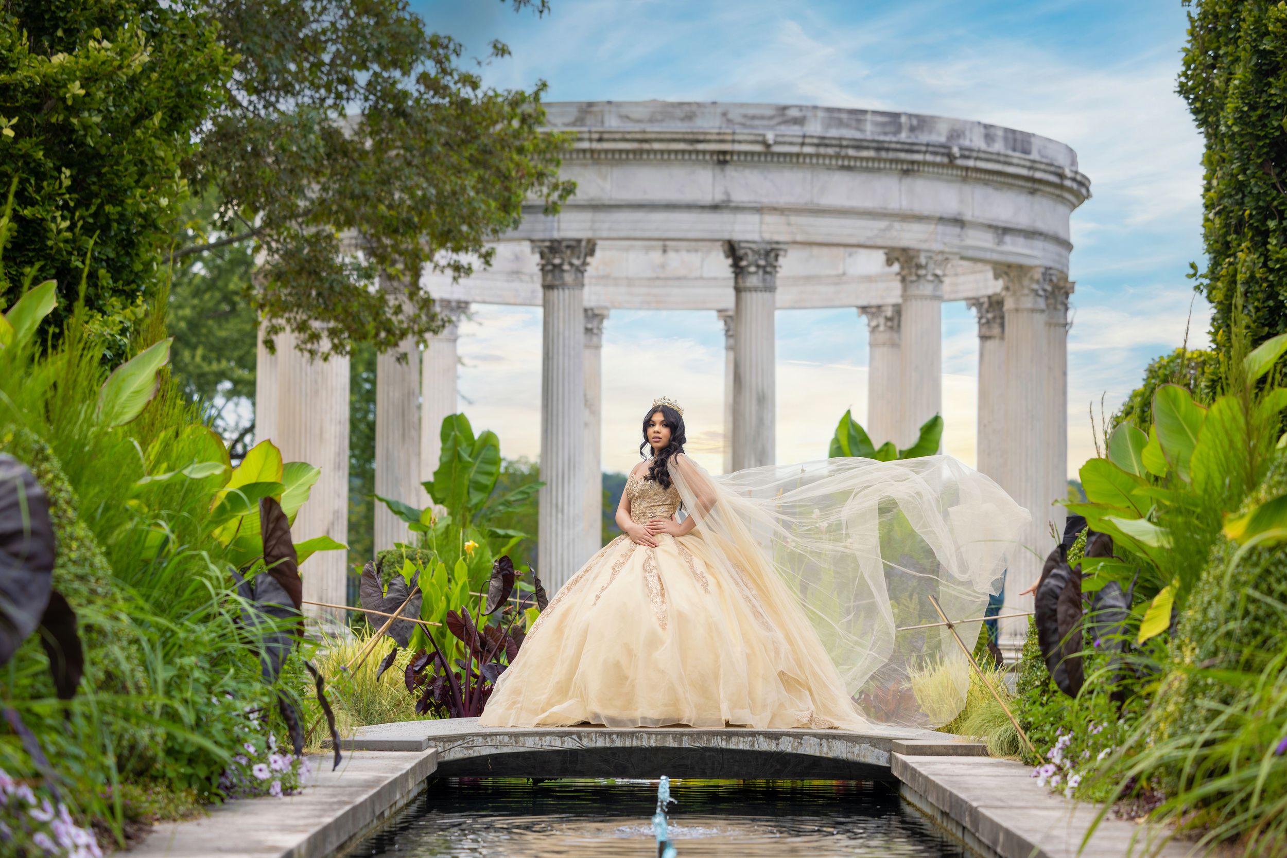 Young woman in a golden ball gown quinceañera dress poses by a fountain with classical columns in a lush garden.