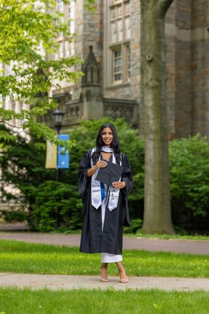 Young woman in black graduation gown and cap poses proudly on a college campus pathway surrounded by green trees.