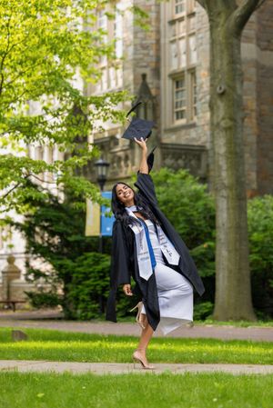 A joyful graduate in black cap and gown tosses her mortarboard in the air on a lush green campus.