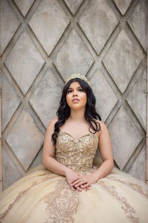Young woman in gold quinceañera gown with lace embroidery and crown poses gracefully against diamond-patterned stone wall.
