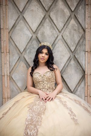 Young woman in gold quinceañera ball gown with crown poses elegantly against diamond-patterned stone wall.