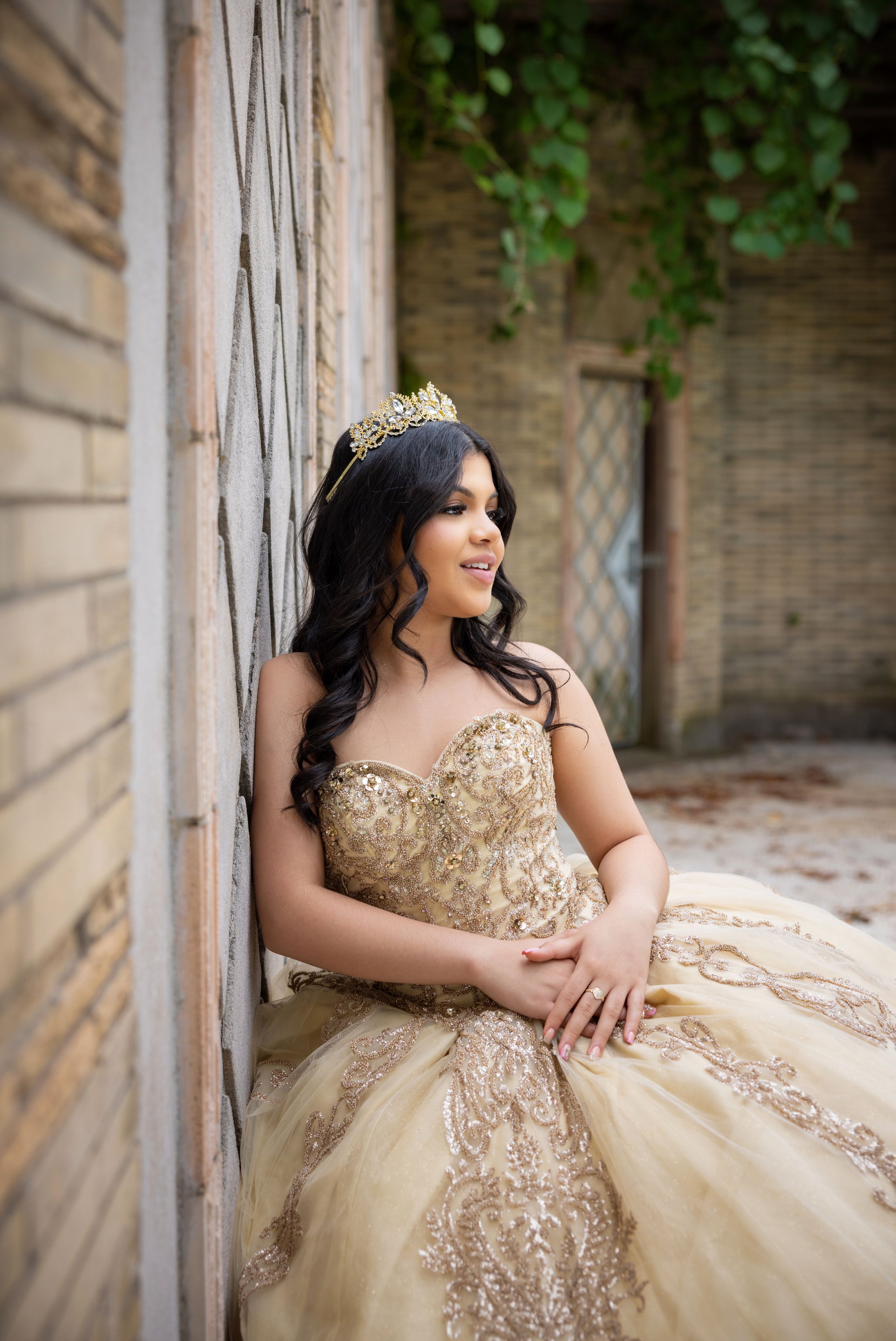 Young woman in gold quinceañera ball gown and tiara crown poses elegantly against rustic brick wall outdoors.