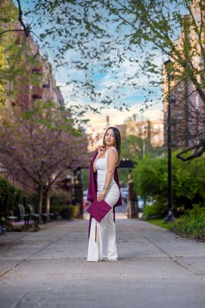 Young woman in white outfit and maroon graduation cap and gown poses on tree-lined path during spring graduation photo shoot.