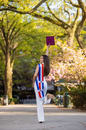 Graduate celebrates outdoors tossing maroon cap wearing Dominican Republic flag outfit among spring trees.
