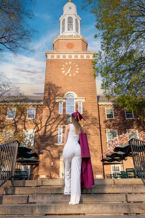 Graduate in white outfit holding maroon cap and gown stands before a brick clock tower on a sunny day.