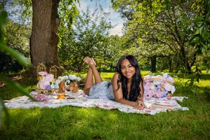 Young woman in floral dress lying on picnic blanket under tree with tea party spread in lush garden.