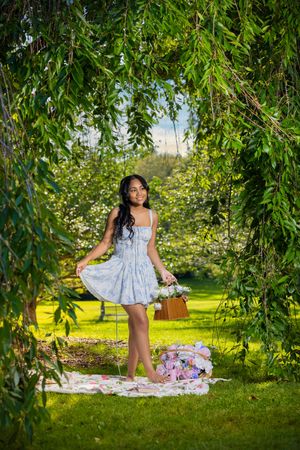 Young woman in light blue dress stands under weeping willow arch on floral blanket in lush green garden.