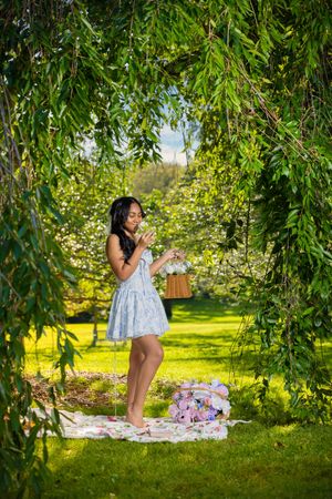 Young woman in blue floral dress holding flower basket under weeping willow tree in lush green garden.