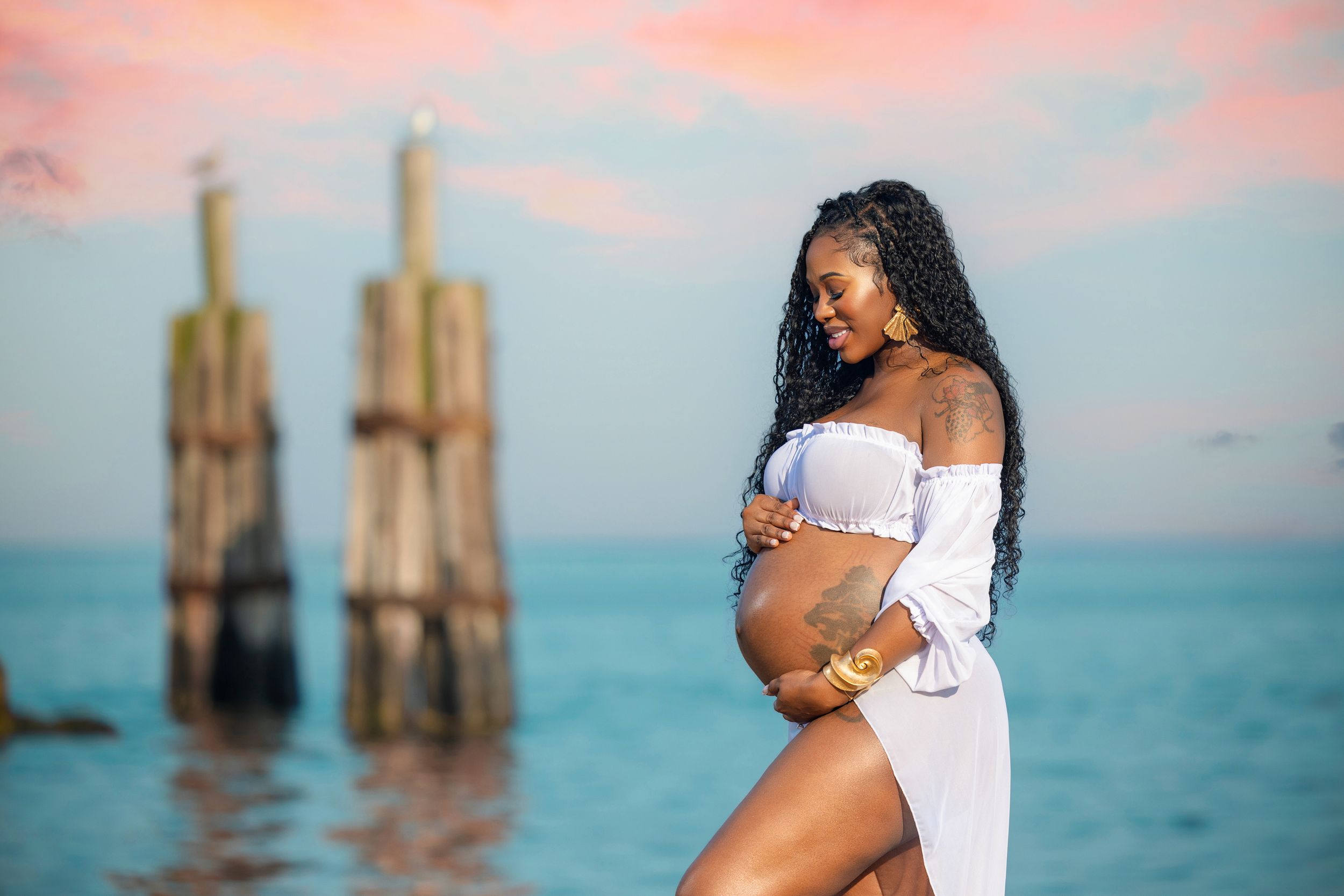Pregnant Black woman in white off-shoulder dress holding baby bump on beach at sunset with wooden pier posts.