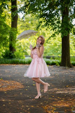 Young woman in pink dress holding lace parasol umbrella, posing in a lush green park pathway.