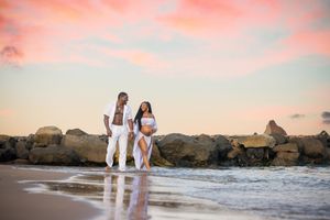 Couple walking on beach at sunset near rocky jetty, wearing white outfits, with pink and blue sky reflecting on wet sand.