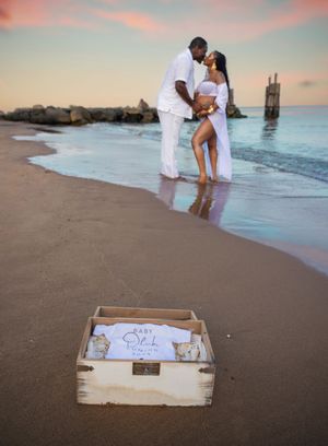 Couple sharing a romantic kiss on a beach at sunset with a baby announcement box in the foreground.