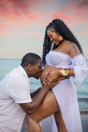 A man kisses a pregnant woman's belly during a romantic maternity photoshoot on the beach at sunset.