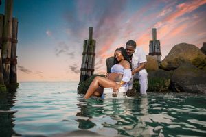 Couple sharing a romantic moment in shallow ocean water at sunset with vibrant pink sky and wooden pier posts behind them.