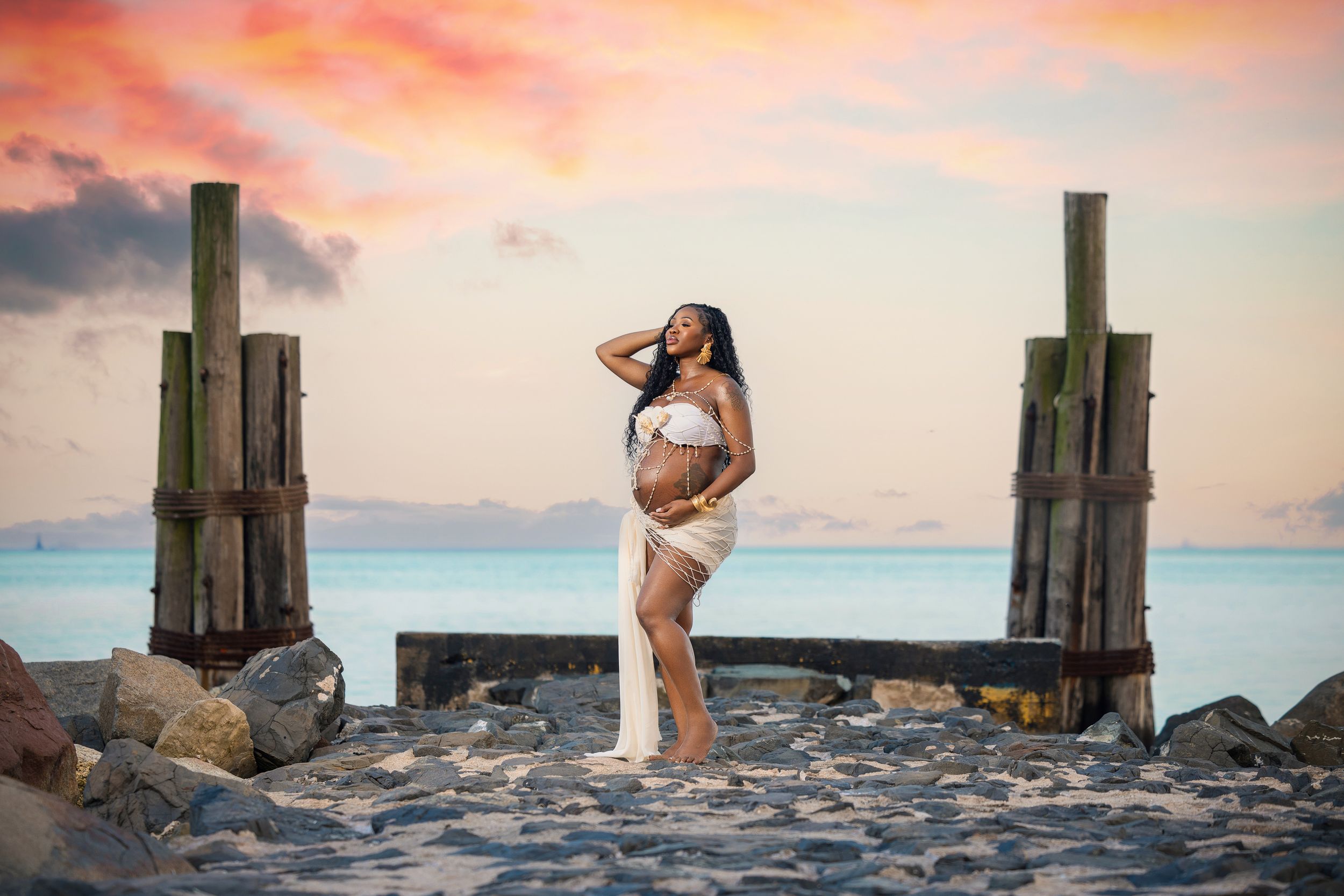 Pregnant woman in white crop top and cream skirt poses on rocky beach at sunset between wooden pier posts.