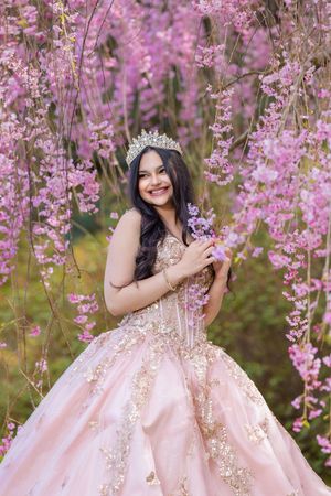 Young woman in pink ball gown and tiara smiles among cascading cherry blossom branches outdoors.