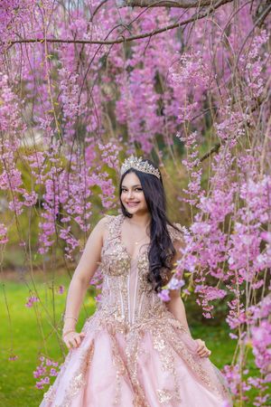 Young woman in pink gold quinceañera gown and tiara smiling among blooming pink cherry blossom trees.