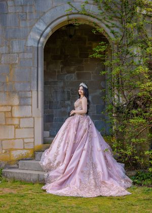 Beautiful woman in ornate pink quinceañera dress and crown standing gracefully in a stone castle doorway.