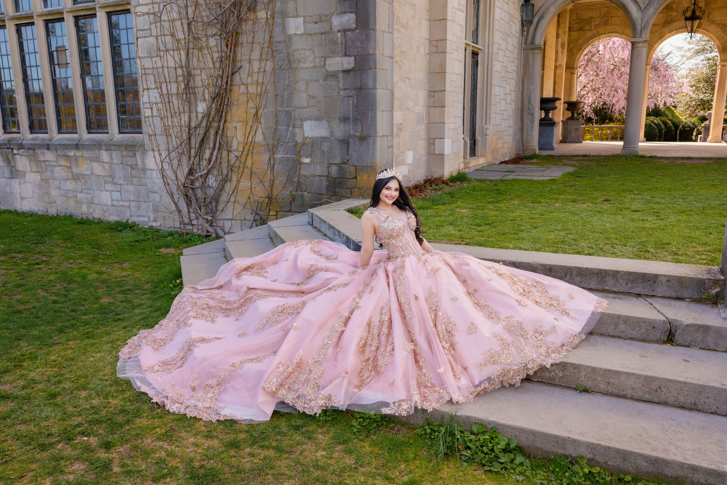 Young woman in a pink ball gown poses gracefully on stone steps outside a castle-like building.