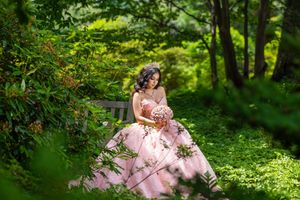 A bride in a pink floral gown sits on a bench surrounded by lush green foliage, holding a bouquet.