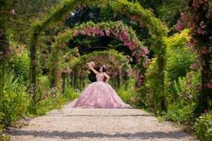 Woman in pink ball gown poses under floral arches in a lush garden pathway blooming with roses.