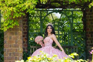 Young woman in pink quinceañera ball gown with floral bouquet and tiara poses in lush garden archway.