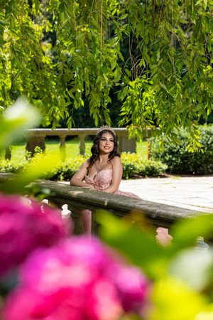 Young woman in pink gown and tiara poses by garden fountain under weeping willow trees with pink flowers.