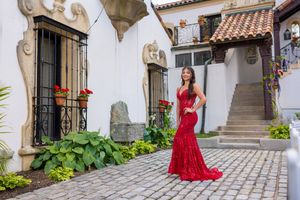 Woman in red lace gown posing in elegant cobblestone courtyard with white walls and terracotta roof.