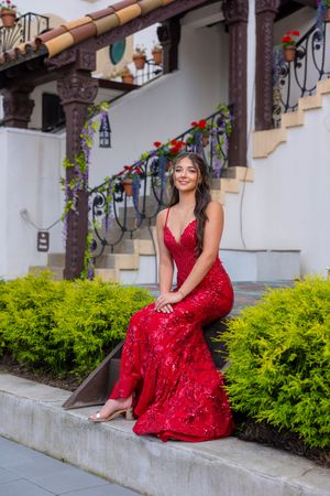 Young woman in elegant red sequin gown sitting gracefully on stone steps surrounded by lush greenery and ornate ironwork.