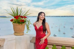 Young woman in red sequin dress poses on waterfront balcony with floral arrangement and sailboats in background.
