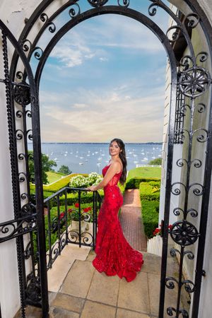 Woman in red sequin gown holding white flowers stands on elegant terrace with wrought iron arch and waterfront view.