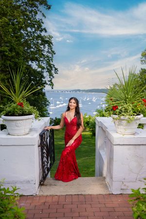 Young woman in red sequin gown poses on garden steps with water and sailboats in the background.