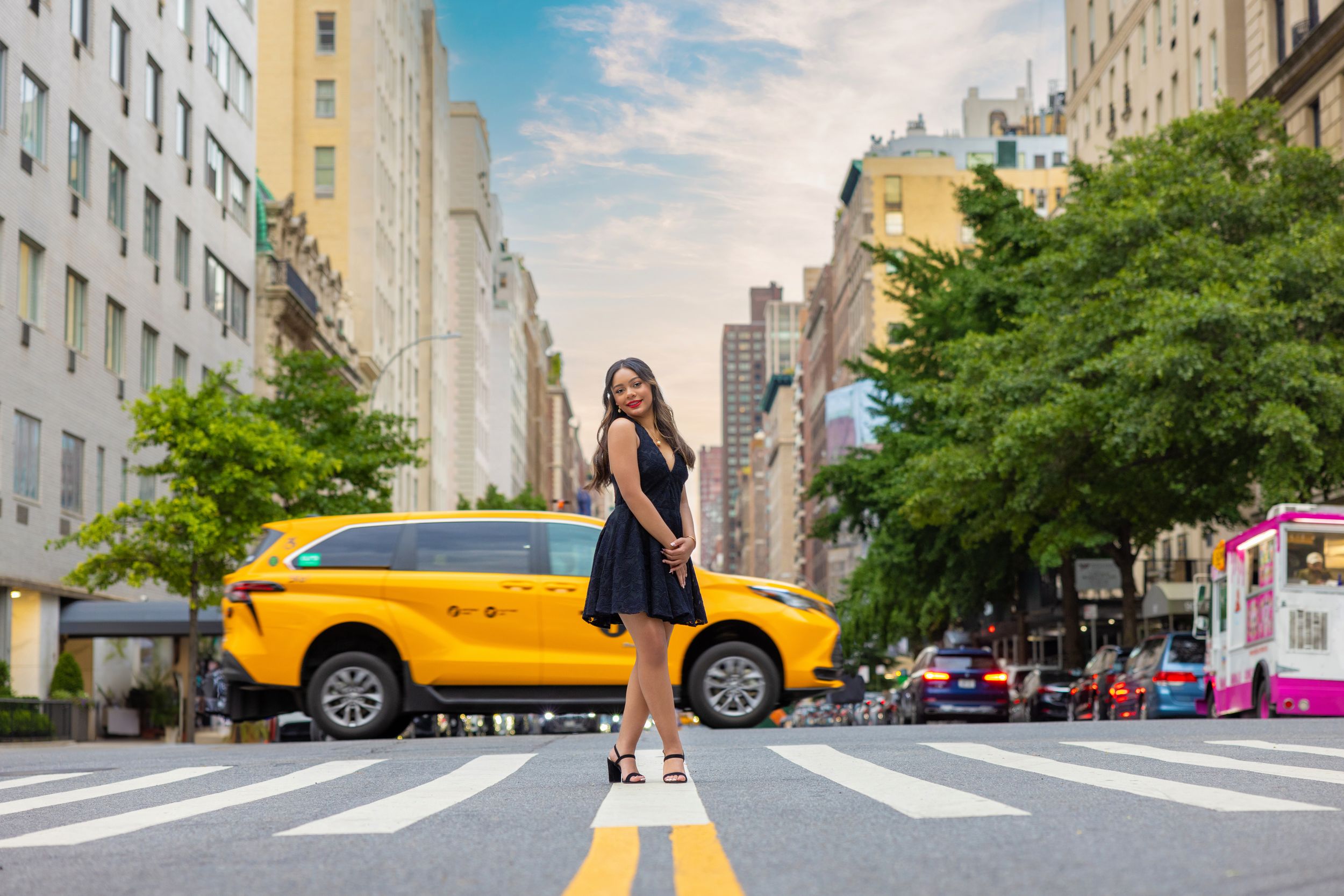 Woman in black dress poses on NYC crosswalk with yellow taxi and tree-lined avenue in background.