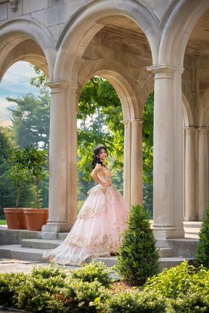 Young woman in pink gold ball gown poses under stone arches surrounded by lush greenery in elegant garden setting.
