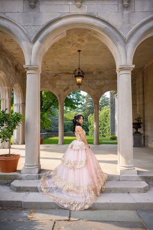 Young woman in pink gold quinceañera ball gown poses gracefully under stone arches of elegant mansion colonnade.