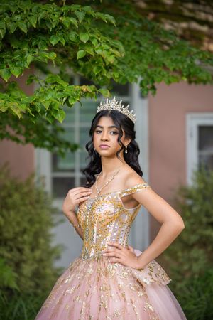 Young woman in pink gold quinceañera gown and tiara crown poses elegantly outdoors near green foliage.