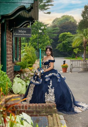 Young woman in navy blue quinceañera ball gown with gold lace appliqués and tiara poses near Main Greenhouse sign.