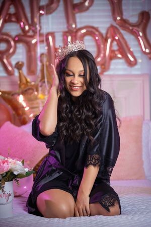 Woman in black satin robe and tiara smiling on bed with pink Happy Birthday balloon decorations.