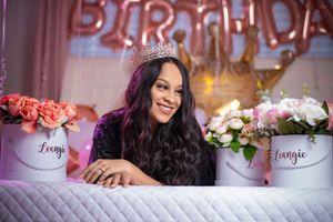 Woman wearing a crown celebrates birthday surrounded by floral arrangements and gold balloon letters spelling BIRTHDAY.