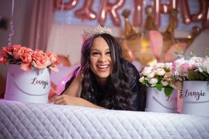 Woman wearing a crown smiles beside floral arrangements labeled Loungie with birthday balloons in background.