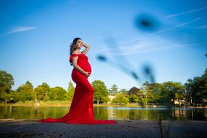 Pregnant woman in elegant red gown posing by a serene lake surrounded by lush green trees under a clear blue sky.