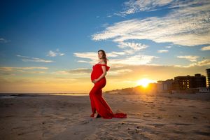 Pregnant woman in red gown poses on beach at sunset, golden light reflecting on sand with ocean and buildings behind.