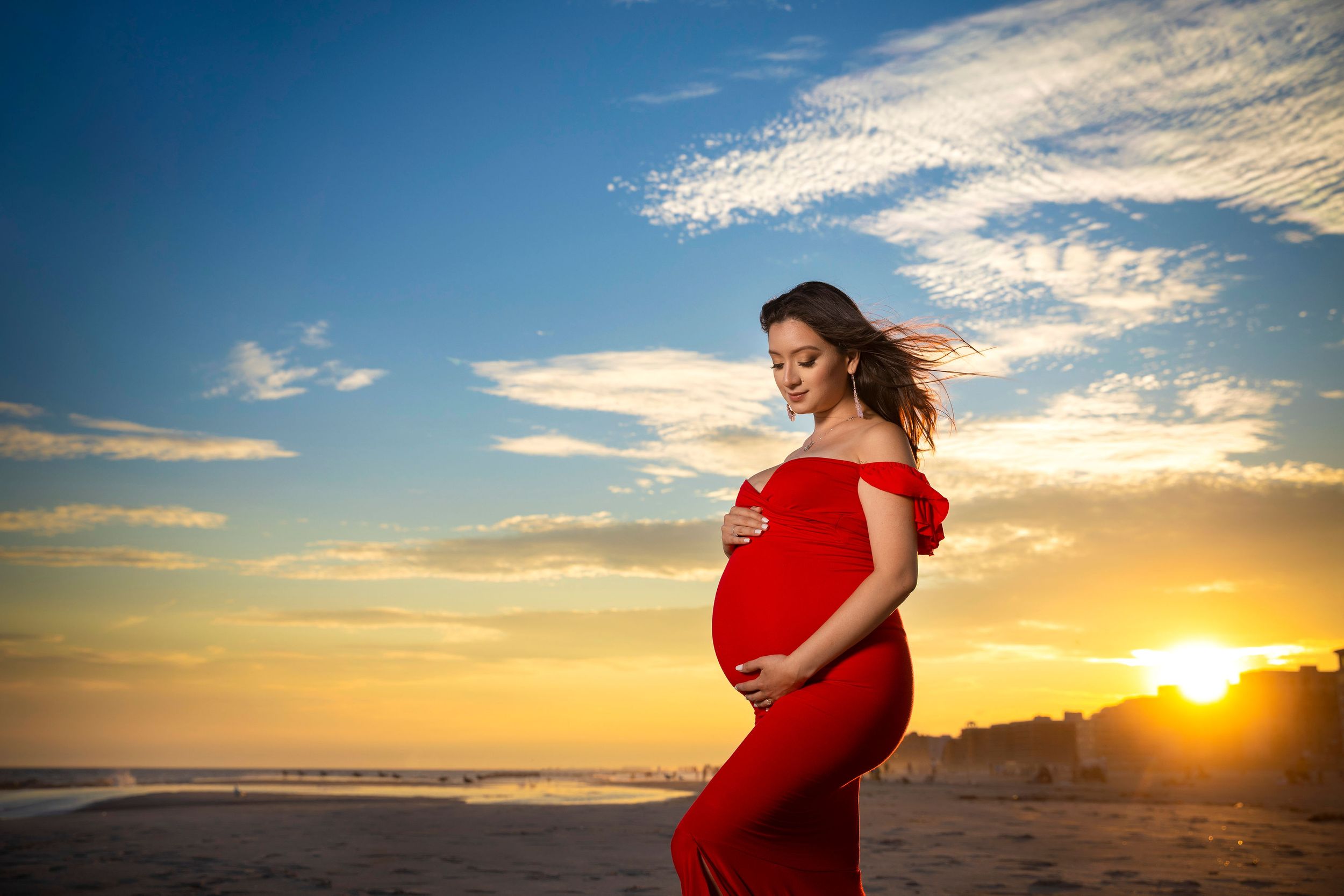 Pregnant woman in red dress on beach at sunset, cradling baby bump with wind in hair and golden sky.