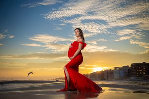Pregnant woman in red gown poses at sunset on beach, golden sky backdrop, maternity photography.